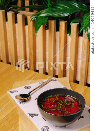Fresh soup served in a black bowl on a wooden table near green plants 140594134