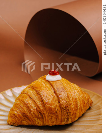 Delicious croissant with cream and cherry on a plate against a brown background Delicious croissant with cream and cherry on a plate against a brown background 140594481