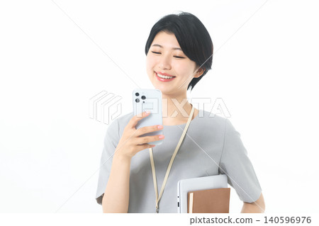 A young woman holding documents and smiling while looking at her smartphone screen against a white background. A young woman holding documents and smiling while looking at her smartphone screen against a white background. 140596976