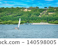 White sailboat sailing on a wide river with lush green hills under blue sky 140598005