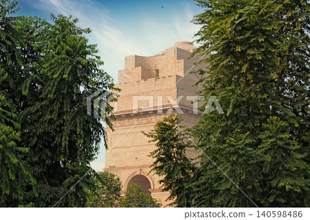 Delhi, India, November 28, 2025: The historic India Gate monument made of sandstone with India inscription viewed through a natural frame of green tree leaves under a blue sky. 140598486