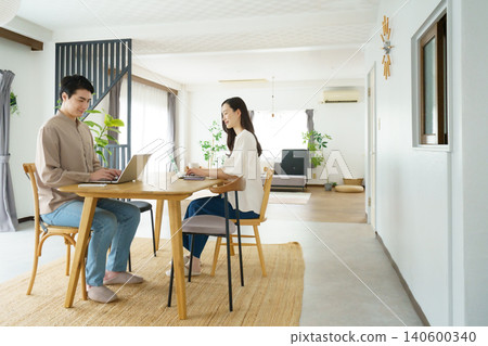 A power couple: a young Japanese man working on his laptop at a table in his home and a young Japanese woman holding a smartphone. A power couple: a young Japanese man working on his laptop at a table in his home and a young Japanese woman holding a smartphone. 140600340