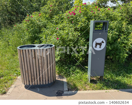 Wooden waste bin, dog waste bag dispenser on park pathway, flowering shrubs. Pet care, urban park infrastructure, responsible dog ownership, public space management, environmental awareness Wooden waste bin, dog waste bag dispenser on park pathway, flowering shrubs. Pet care, urban park infrastructure, responsible dog ownership, public space management, environmental awareness 140602634