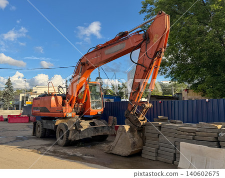 Orange excavator stands at construction site in Moscow beside stacked paving stones. Construction industry marketing, infrastructure development, engineering, heavy equipment Orange excavator stands at construction site in Moscow beside stacked paving stones. Construction industry marketing, infrastructure development, engineering, heavy equipment 140602675