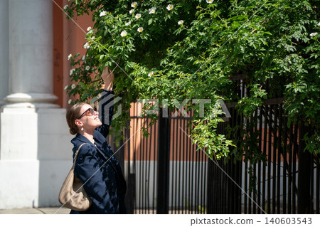 Young woman in dark blazer and sunglasses reaches toward blooming tree branch on sunny city street surrounded by lush green foliage. Spring season, urban nature advertising, outdoor leisure Young woman in dark blazer and sunglasses reaches toward blooming tree branch on sunny city street surrounded by lush green foliage. Spring season, urban nature advertising, outdoor leisure 140603543