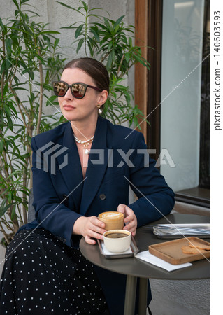 Young woman in sunglasses enjoys coffee at outdoor cafe table surrounded by green plants in modern urban setting. Leisure content, social media, hospitality promotion, urban relaxation branding Young woman in sunglasses enjoys coffee at outdoor cafe table surrounded by green plants in modern urban setting. Leisure content, social media, hospitality promotion, urban relaxation branding 140603593