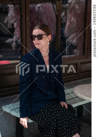 Young woman in sunglasses sits on outdoor bench with sparkling disco ball reflections across clothing and skin against stylish cafe backdrop. Lifestyle, creative lighting, fashion Young woman in sunglasses sits on outdoor bench with sparkling disco ball reflections across clothing and skin against stylish cafe backdrop. Lifestyle, creative lighting, fashion 140603598