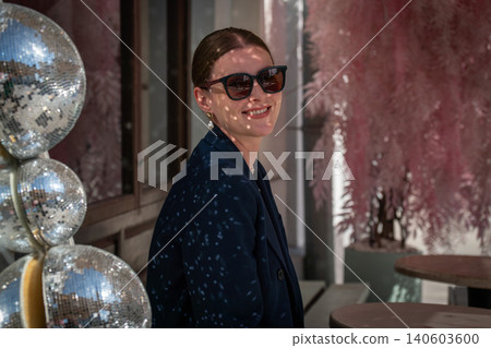 Smiling young woman in sunglasses sits beside mirrored disco balls with sparkling light reflections across blazer, face in stylish outdoor cafe setting. Lifestyle, creative lighting, cafe culture Smiling young woman in sunglasses sits beside mirrored disco balls with sparkling light reflections across blazer, face in stylish outdoor cafe setting. Lifestyle, creative lighting, cafe culture 140603600