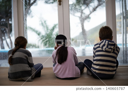 Rain, window and sad child on a floor unhappy, upset or annoyed by bad weather at home. Glass, disappointed and sibling kids in a living room frustrated by storm, cold or unexpected winter disaster 140603787