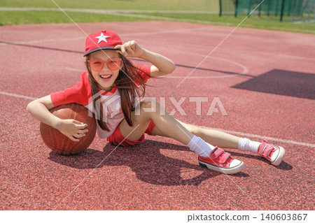 A laughing girl in a cap and red glasses sits on a basketball court. Sports education for children. A laughing girl in a cap and red glasses sits on a basketball court. Sports education for children. 140603867
