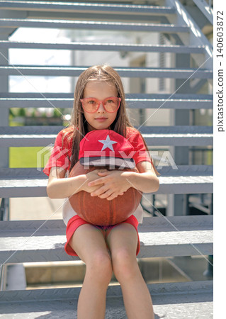 A cute, beautiful athlete girl is sitting on the stairs hugging a basketball covered with a cap. 140603872