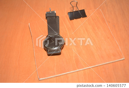 Black analog wristwatch and a single metal binder on a wooden desk with fastener Black analog wristwatch and a single metal binder on a wooden desk with fastener 140605577