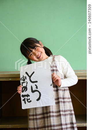 A female elementary school student smiles and shows her calligraphy artwork of a tiger at the back of the classroom. 140607309