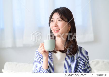 一位面帶微笑的日本女性坐在沙發上喝咖啡。 一位面帶微笑的日本女性坐在沙發上喝咖啡。 140608031