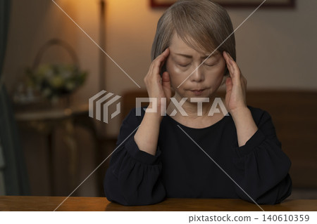 A senior woman is holding her head in her hands in her living room, feeling unwell. 140610359