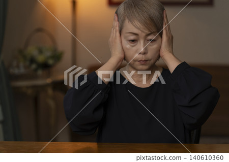 一位老婦人雙手抱頭坐在客廳裡，感覺身體不適。 140610360