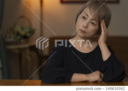 A senior woman is holding her head in her hands in her living room, feeling unwell. 140610362