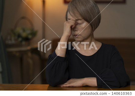 A senior woman is holding her head in her hands in her living room, feeling unwell. 140610363