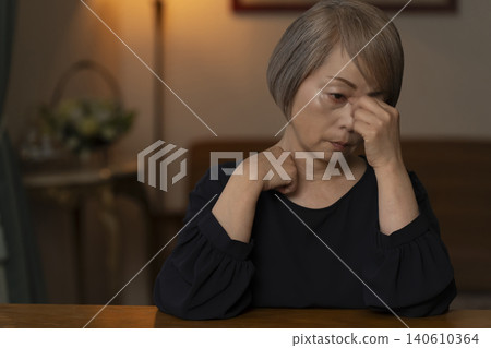 一位老婦人雙手抱頭坐在客廳裡,感覺身體不適。 一位老婦人雙手抱頭坐在客廳裡,感覺身體不適。 140610364