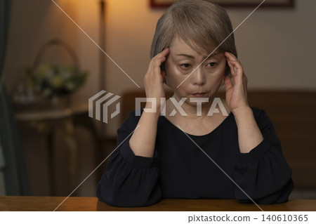 一位老婦人雙手抱頭坐在客廳裡，感覺身體不適。 140610365