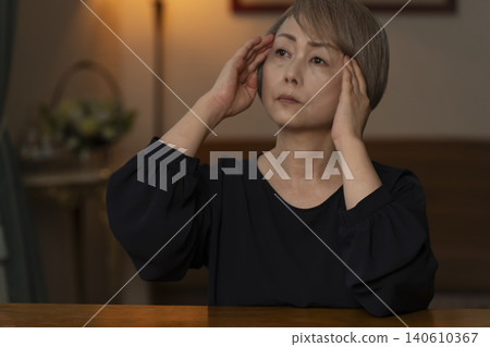 一位老婦人雙手抱頭坐在客廳裡，感覺身體不適。 140610367