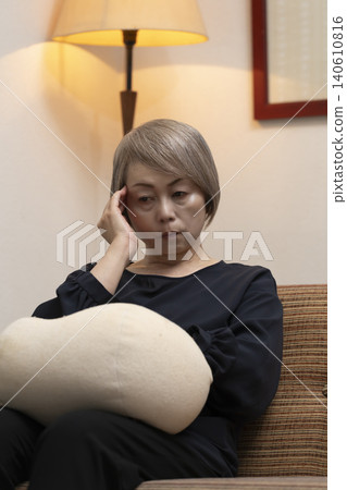 A senior woman is sitting on the living room sofa, holding her head in her hands due to feeling unwell. A senior woman is sitting on the living room sofa, holding her head in her hands due to feeling unwell. 140610816