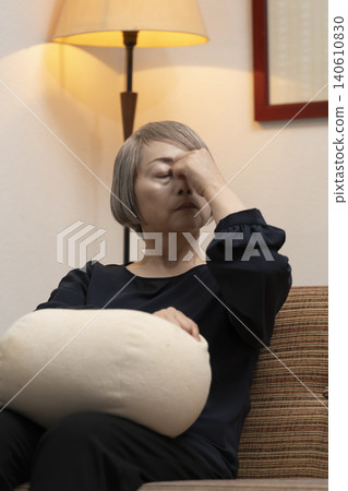 A senior woman is sitting on the living room sofa, holding her head in her hands due to feeling unwell. 140610830