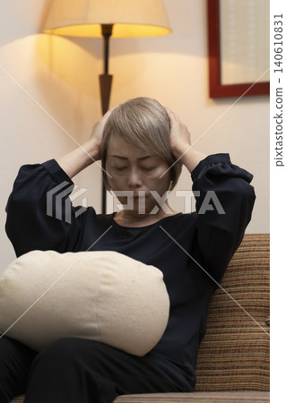 A senior woman is sitting on the living room sofa, holding her head in her hands due to feeling unwell. 140610831