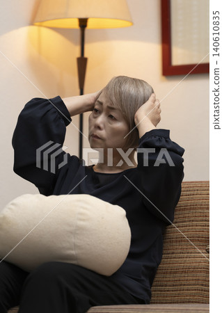 一位老婦人坐在客廳沙發上，雙手抱頭，似乎身體不適。 140610835