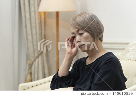 一位老婦人坐在客廳沙發上,雙手抱頭,似乎身體不適。 一位老婦人坐在客廳沙發上,雙手抱頭,似乎身體不適。 140610839