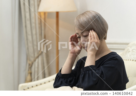 A senior woman is sitting on the living room sofa, holding her head in her hands due to feeling unwell. A senior woman is sitting on the living room sofa, holding her head in her hands due to feeling unwell. 140610842
