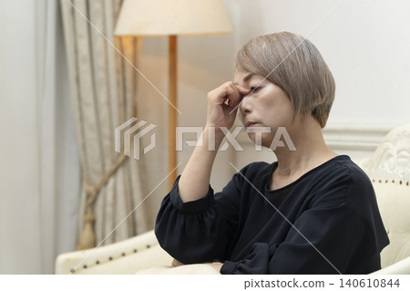 一位老婦人坐在客廳沙發上,雙手抱頭,似乎身體不適。 一位老婦人坐在客廳沙發上,雙手抱頭,似乎身體不適。 140610844