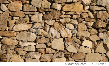 A wall of rough natural stone laid using the dry-laying method. Stones of various sizes and shapes. The surface of the stones is rough, textured, with natural chips, cracks, and irregularities. A wall of rough natural stone laid using the dry-laying method. Stones of various sizes and shapes. The surface of the stones is rough, textured, with natural chips, cracks, and irregularities. 140617943