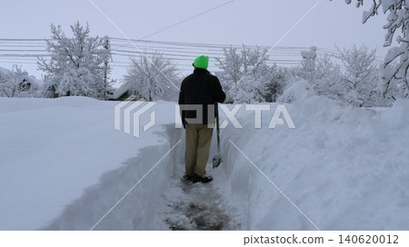 A man clearing deep snowdrifts after a heavy snowfall in a rural area or private property. The man is inside a deep, narrow trench he has cut in the snow 140620012
