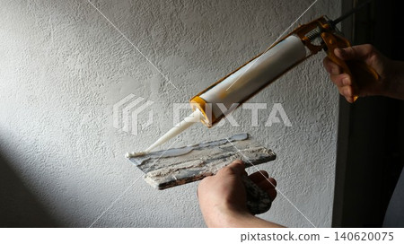 The process of applying mortar or sealant during renovation work. A man holds a yellow skeleton gun with a filled white tube and a wide metal spatula covered in dried remains of old putty or plaster. 140620075