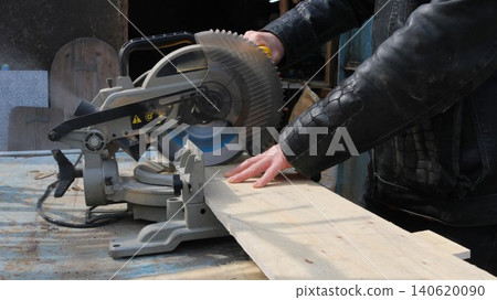 The miter saw is mounted on a workbench. The saw blade is partially lowered and covered by a protective guard. Yellow controls and safety warning signs are visible on the housing. A man in a black jac 140620090