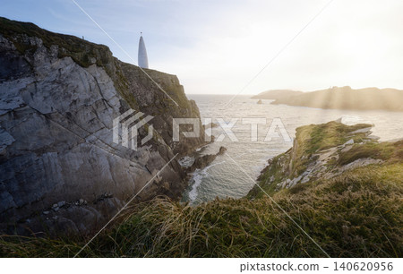 A tall, conical white structure stands on a rocky cliff overlooking the ocean with sunbeams breaking through the sky. A tall, conical white structure stands on a rocky cliff overlooking the ocean with sunbeams breaking through the sky. 140620956