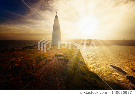 A conical tower stands on a grassy cliff overlooking the ocean during a bright sunset with sunbeams shining on the water. A conical tower stands on a grassy cliff overlooking the ocean during a bright sunset with sunbeams shining on the water. 140620957