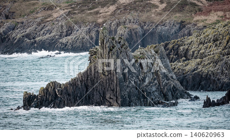 Jagged rock formations rise from the ocean, with waves crashing against their base and a dry, grassy hillside in the background. Jagged rock formations rise from the ocean, with waves crashing against their base and a dry, grassy hillside in the background. 140620963
