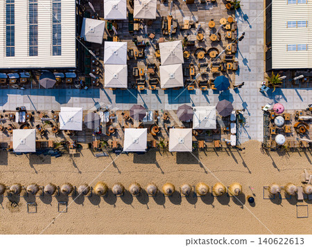 Aerial top-down view of a luxury beach club terrace with wooden furniture, large sun umbrellas, and straw parasols on the sandy shore of Scheveningen, Netherlands. Aerial top-down view of a luxury beach club terrace with wooden furniture, large sun umbrellas, and straw parasols on the sandy shore of Scheveningen, Netherlands. 140622613