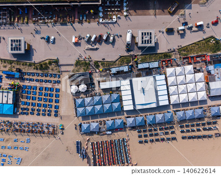 Aerial top-down view of coastal beach clubs, colorful sun umbrellas, sandy shores, and retro camper vans parked along the promenade in Scheveningen, Netherlands. Aerial top-down view of coastal beach clubs, colorful sun umbrellas, sandy shores, and retro camper vans parked along the promenade in Scheveningen, Netherlands. 140622614