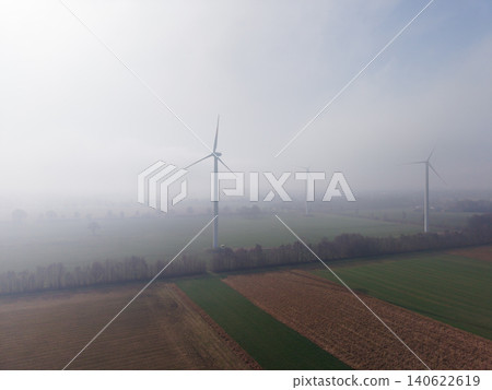 Aerial view of industrial wind turbines standing in misty green and brown agricultural fields under bright morning sun fog in the Dutch countryside, Netherlands. 140622619