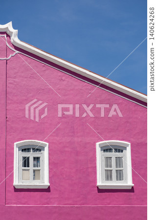 Bright pink wall of a building featuring two white-framed windows under a clear blue sky, showcasing vibrant color contrast and architectural details in a sunny environment Bright pink wall of a building featuring two white-framed windows under a clear blue sky, showcasing vibrant color contrast and architectural details in a sunny environment 140624268