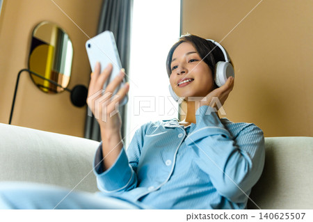 A woman sitting on a sofa with headphones on, looking at her smartphone. A woman sitting on a sofa with headphones on, looking at her smartphone. 140625507