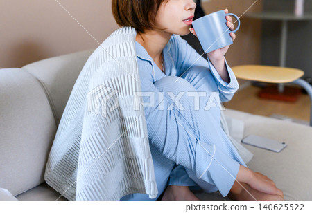 A woman wrapped in a blanket on a sofa, drinking coffee. A woman wrapped in a blanket on a sofa, drinking coffee. 140625522