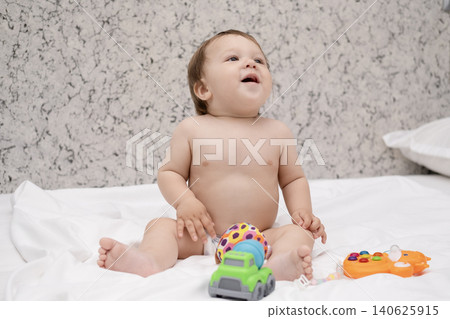 Adorable diaper baby boy sitting on white bed and reaching for toy Adorable diaper baby boy sitting on white bed and reaching for toy 140625915