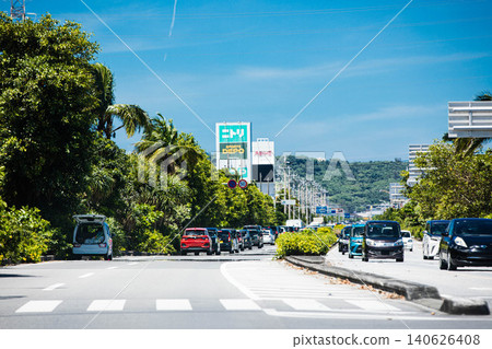 Roadside scenery around Maehara, Uruma City, Okinawa Prefecture: A main road lined with palm trees against a blue sky. 140626408