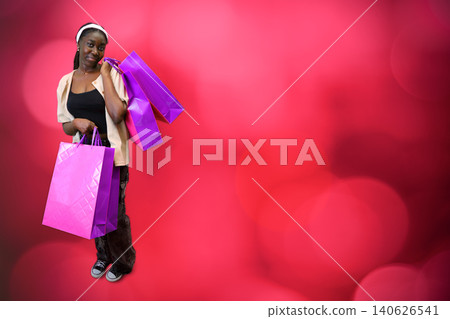 Smiling young Black woman holding purple shopping bags against a bright pink bokeh background with copy space 140626541