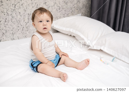 Cute baby boy in tank top sitting on bed and scratching his head thoughtfully Cute baby boy in tank top sitting on bed and scratching his head thoughtfully 140627097