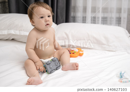 Adorable diaper baby boy sitting on white bed and reaching for toy Adorable diaper baby boy sitting on white bed and reaching for toy 140627103